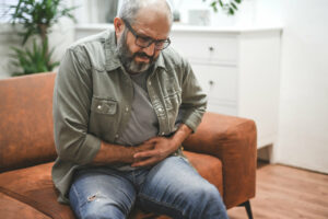 A mature adult man is experiencing stomach pain due to acid reflux while sitting on his couch.