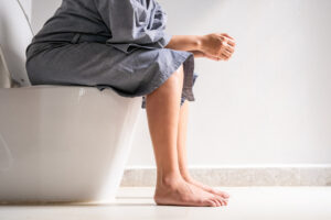 A woman sits on the toilet bowl in the bathroom, experiencing stomach pain and digestive issues due to Crohn's disease.